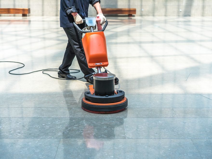 A worker steering an orange floor scrubbing machine across a polished tiled floor