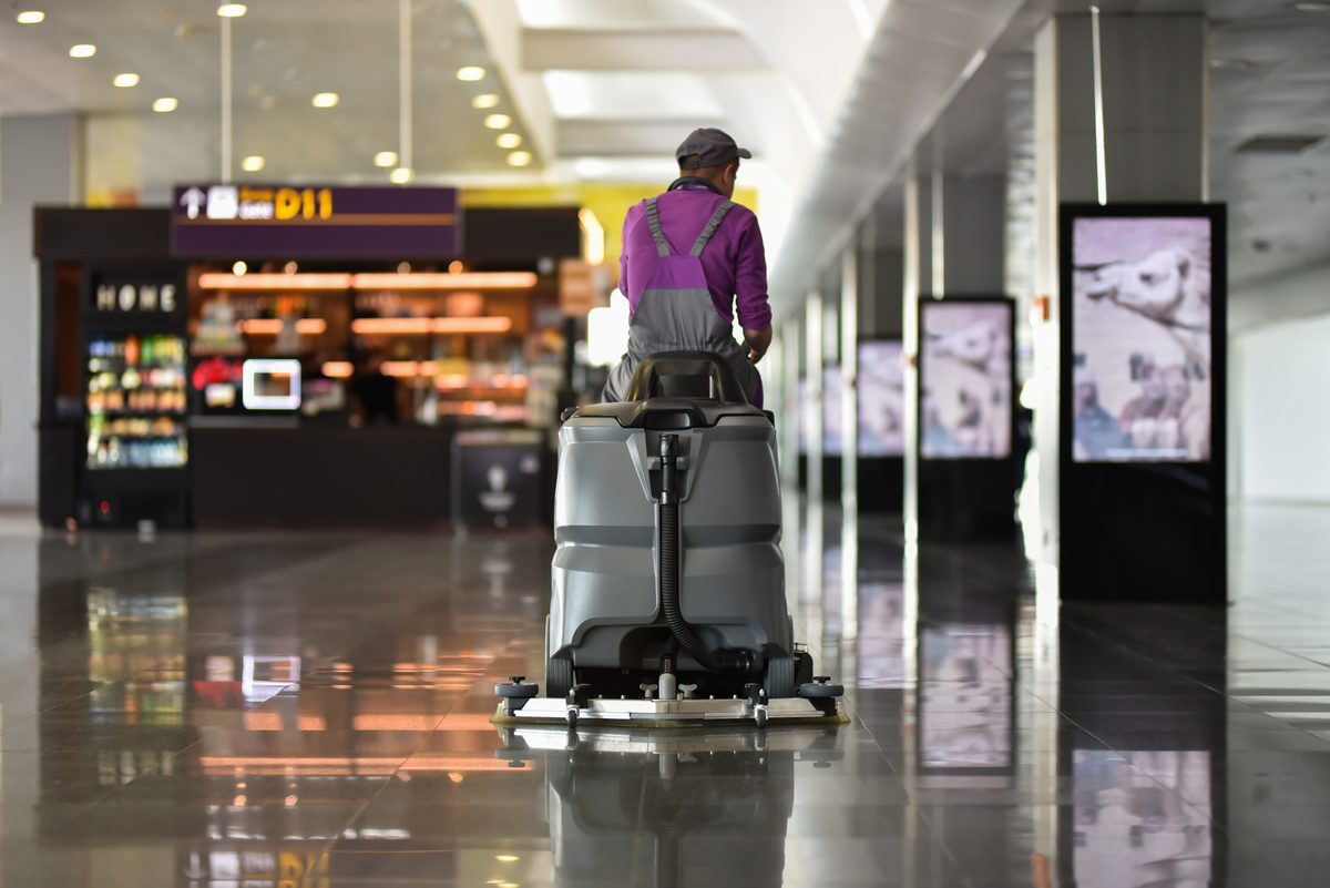 An operator on a ride-on floor scrubber working along the polished floor of a shopping centre concourse
