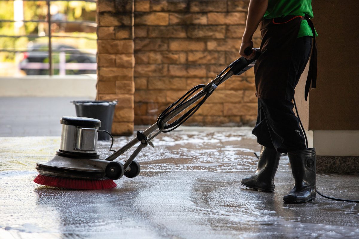 A worker in rubber boots using a floor scrubbing machine on a wet floor outside a brick building