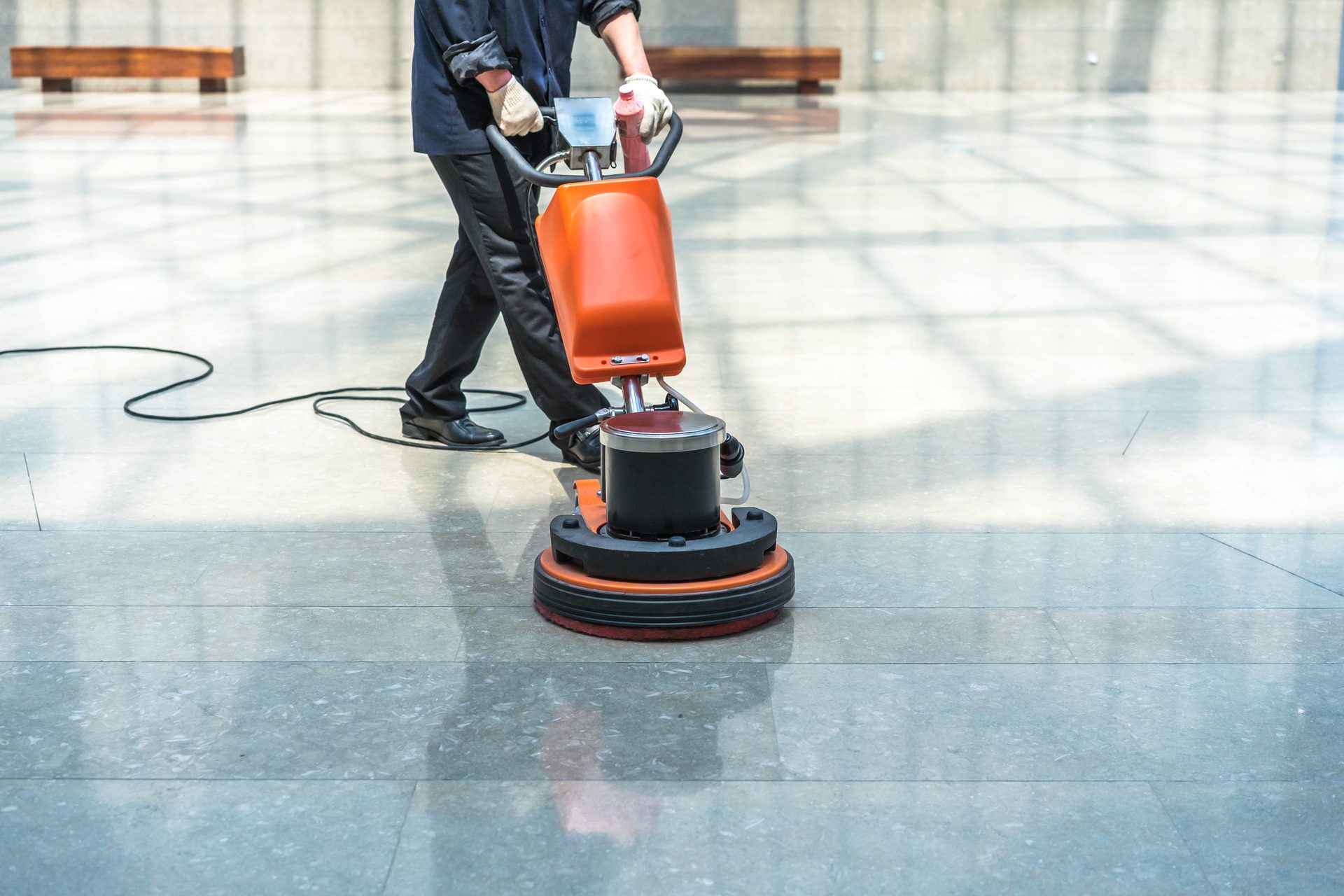 A worker steering an orange floor scrubbing machine across a polished tiled floor