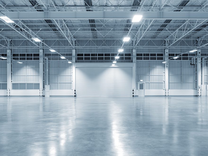 An empty warehouse with a polished grey concrete floor under rows of overhead lights, a roller door at the far end